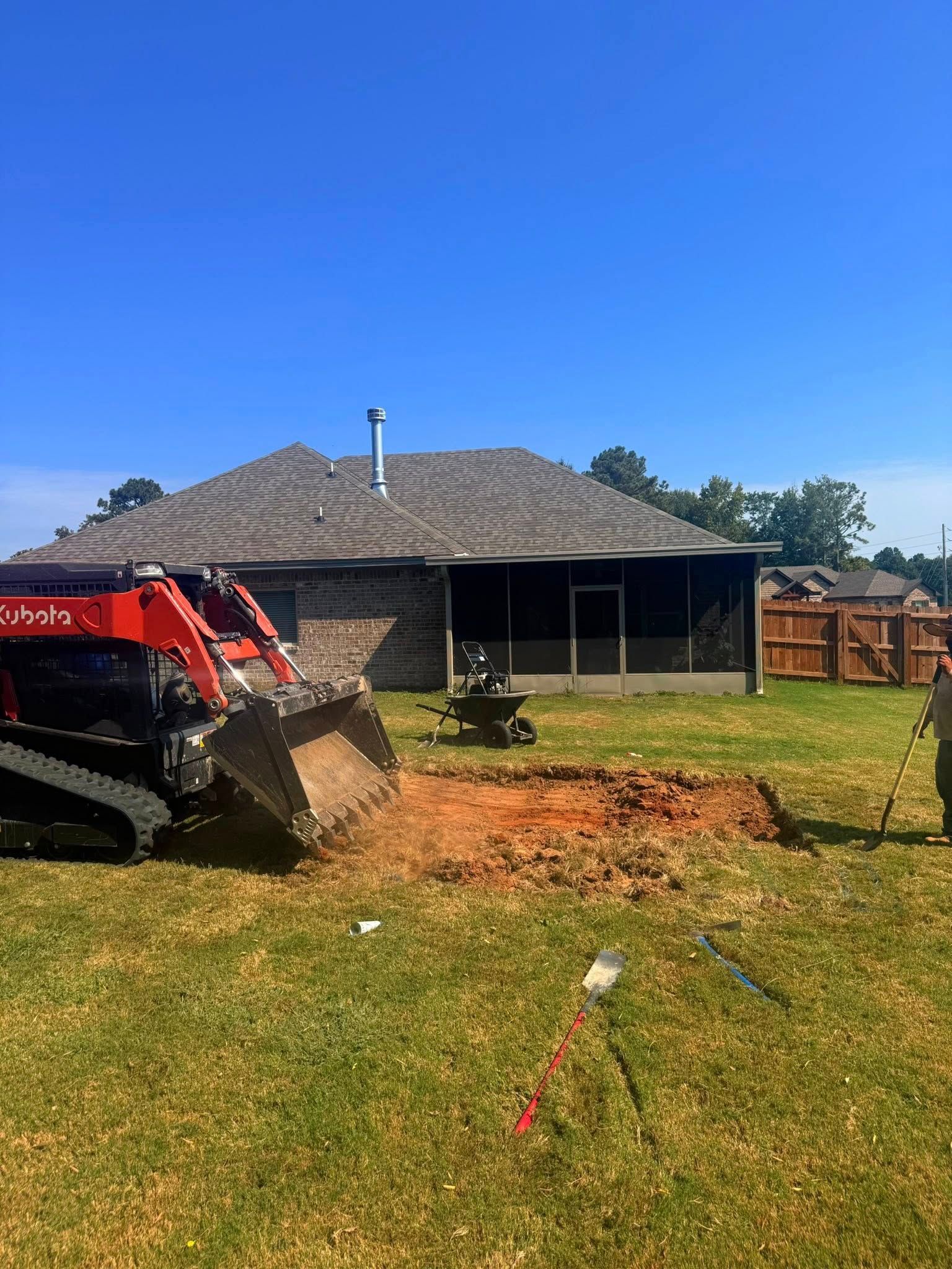 A small excavator digs in a backyard, near a house with a screened porch and a blue sky.