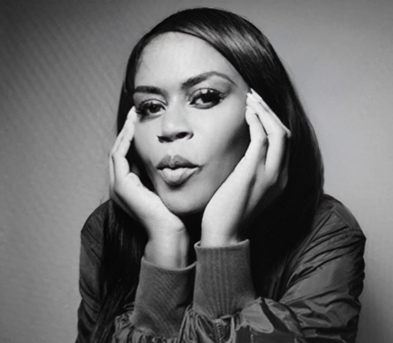 Black-and-white portrait of a person resting their face in both hands, looking at the camera.