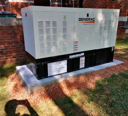 A Generac generator on a concrete pad near a brick building, surrounded by wood chips.