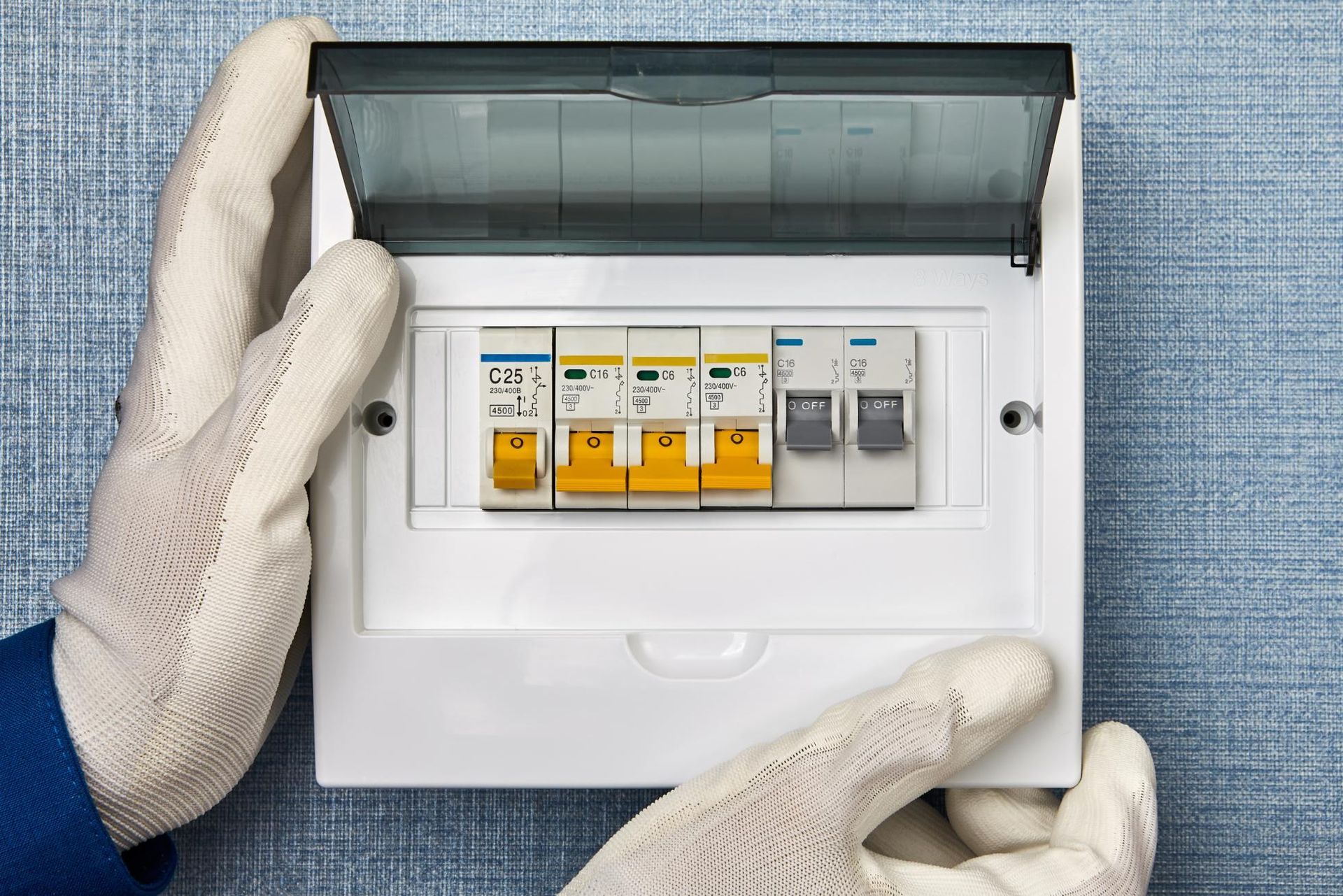 Hands in white gloves open a white electrical breaker box with colorful switches, mounted on a blue wall.
