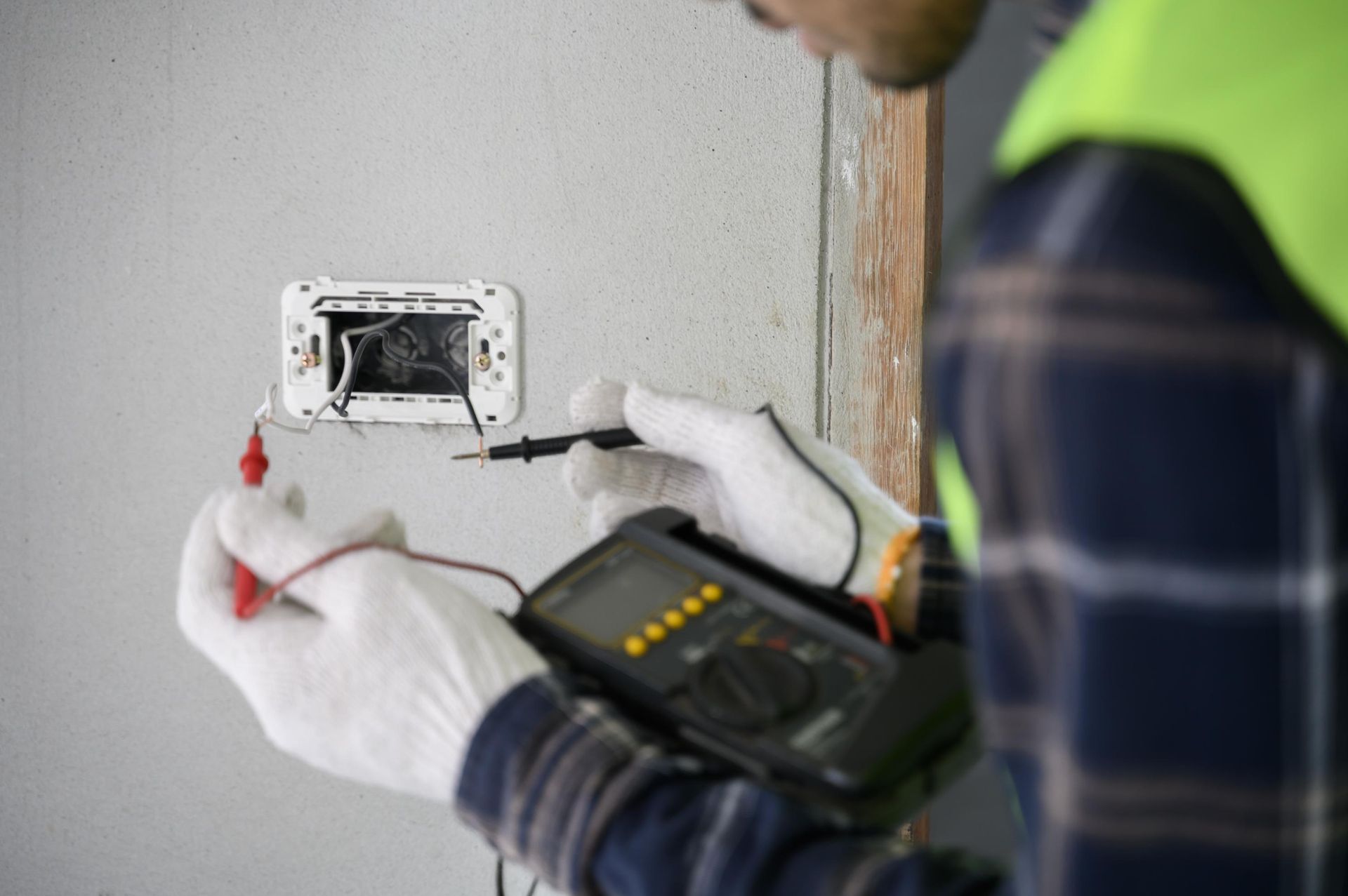 Electrician tests exposed wires in a wall outlet with a multimeter, wearing gloves.