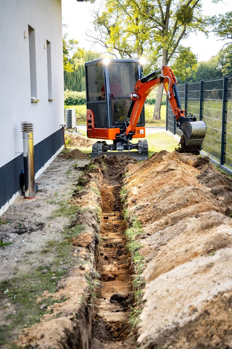 Mini excavator digging a trench beside a building. Orange and black machine, dirt, grass, sunny day. Mini excavator digging a trench beside a building. Orange and black machine, dirt, grass, sunny day.