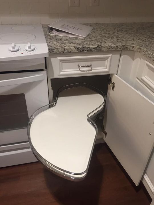 A white cabinet base with an open, kidney-shaped pull-out shelf next to a white oven and granite countertop.
