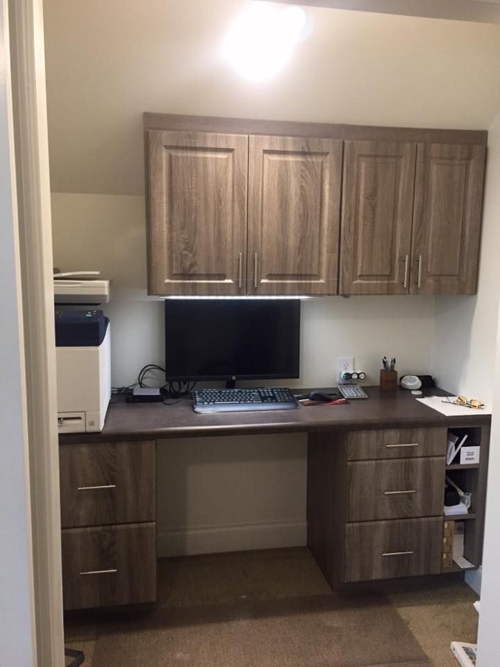 A built-in wood-toned desk and cabinet unit in a small alcove, featuring a computer monitor, keyboard, and printer.