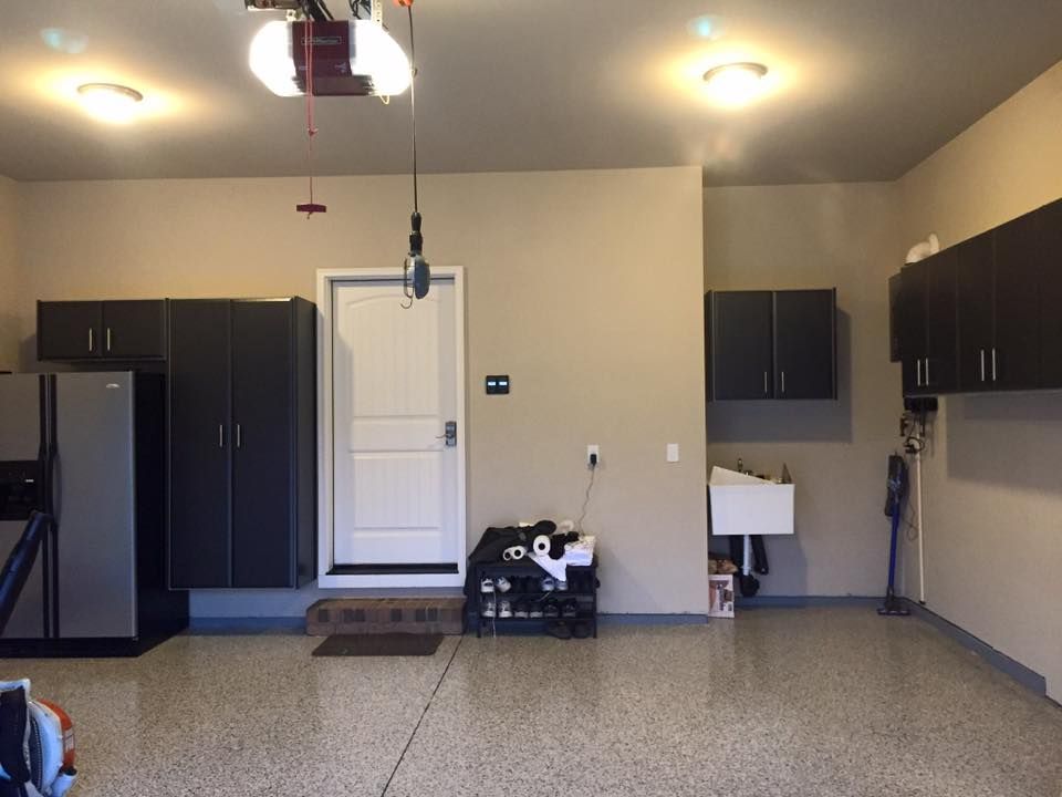 A brightly lit garage with grey epoxy flooring, dark cabinetry, a white utility sink, and a door leading outside.