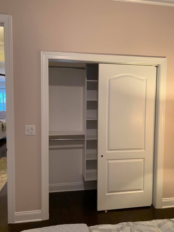 A white closet with an open door, revealing a hanging rod and built-in shelving inside against a light pink wall.