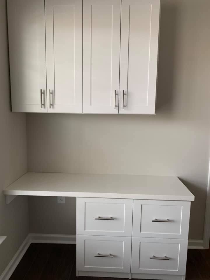 White built-in desk with overhead cabinets and four drawers against a light wall over dark wood flooring.