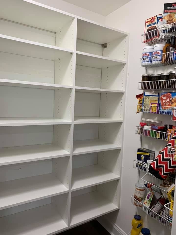 A white walk-in pantry with two vertical shelving units and a wire rack on the door filled with various food items.