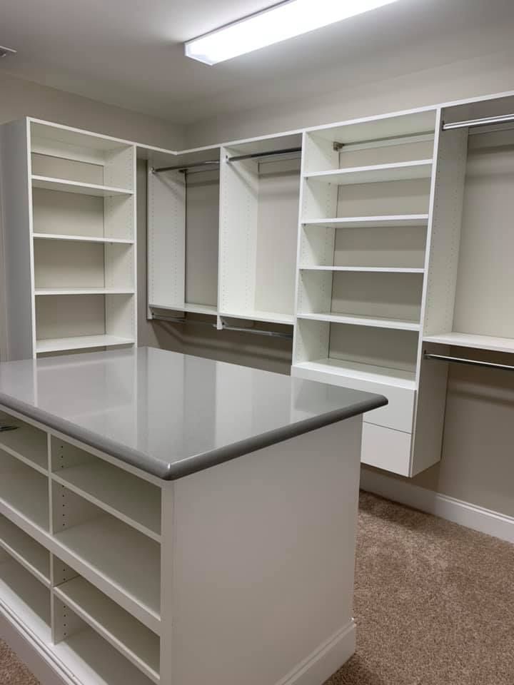A white walk-in closet featuring floor-to-ceiling shelving, hanging rods, and a central island with a grey countertop.