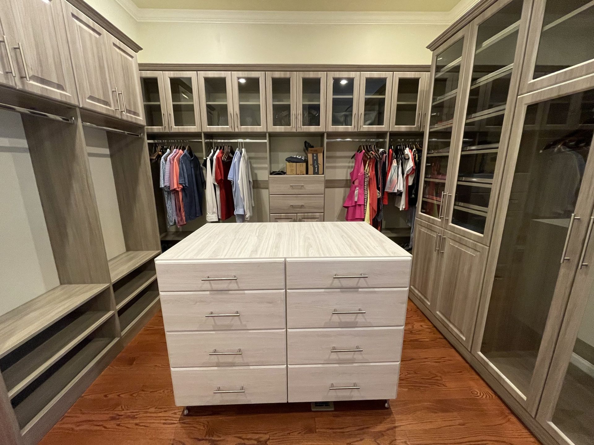 A walk-in closet with light wood cabinetry, hanging clothes, glass-front cabinets, and a central dresser with eight drawers.
