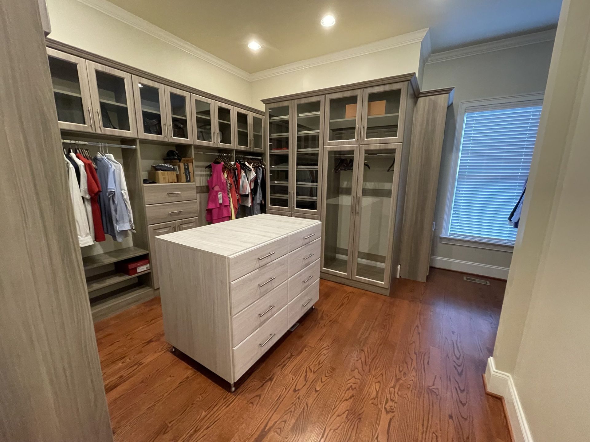 A walk-in closet featuring light wood cabinetry with glass doors, a central storage island with drawers, and wood flooring.