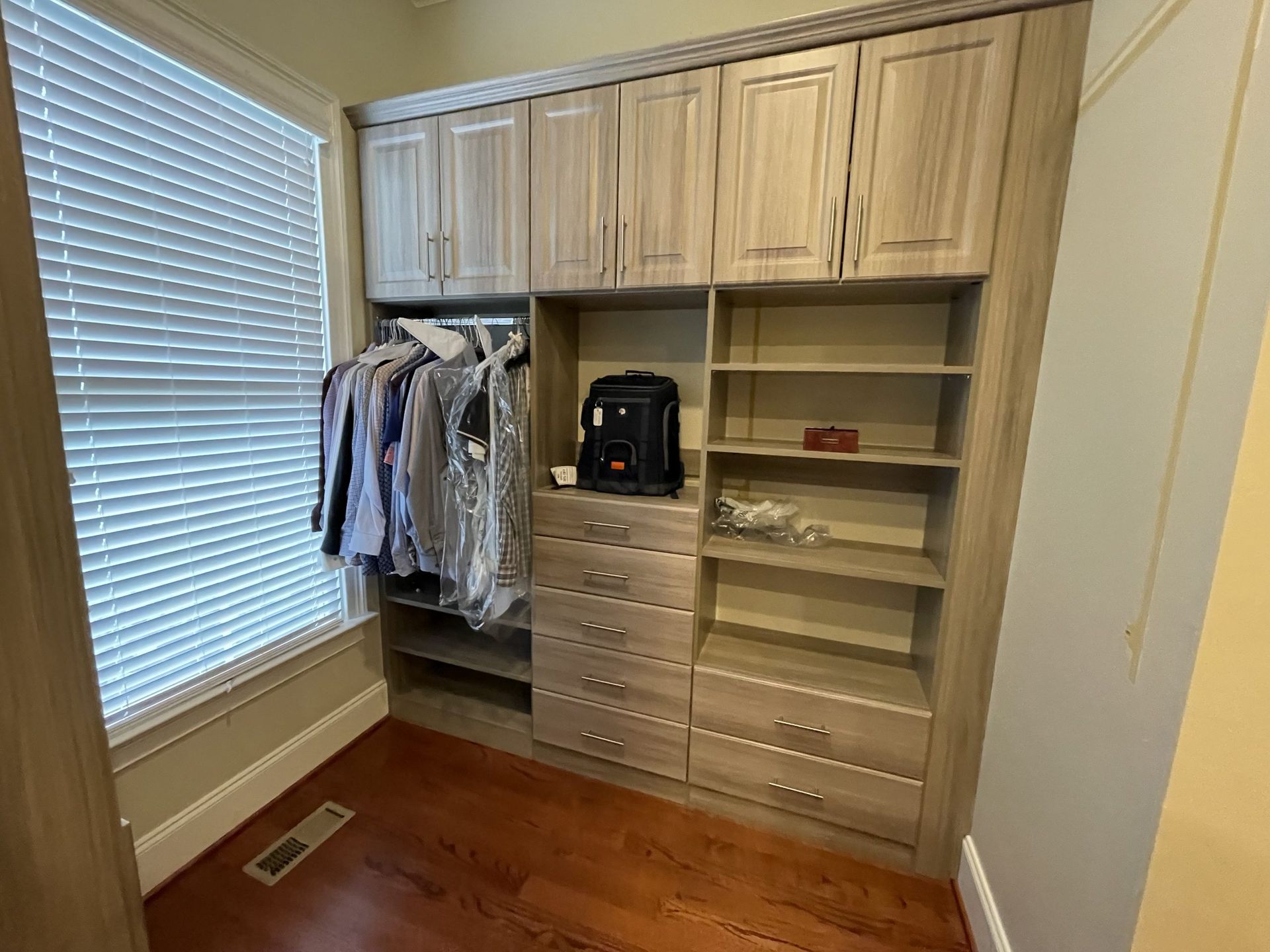 Light wood custom closet unit with hanging space, open shelving, and a column of drawers in a room with hardwood floors.