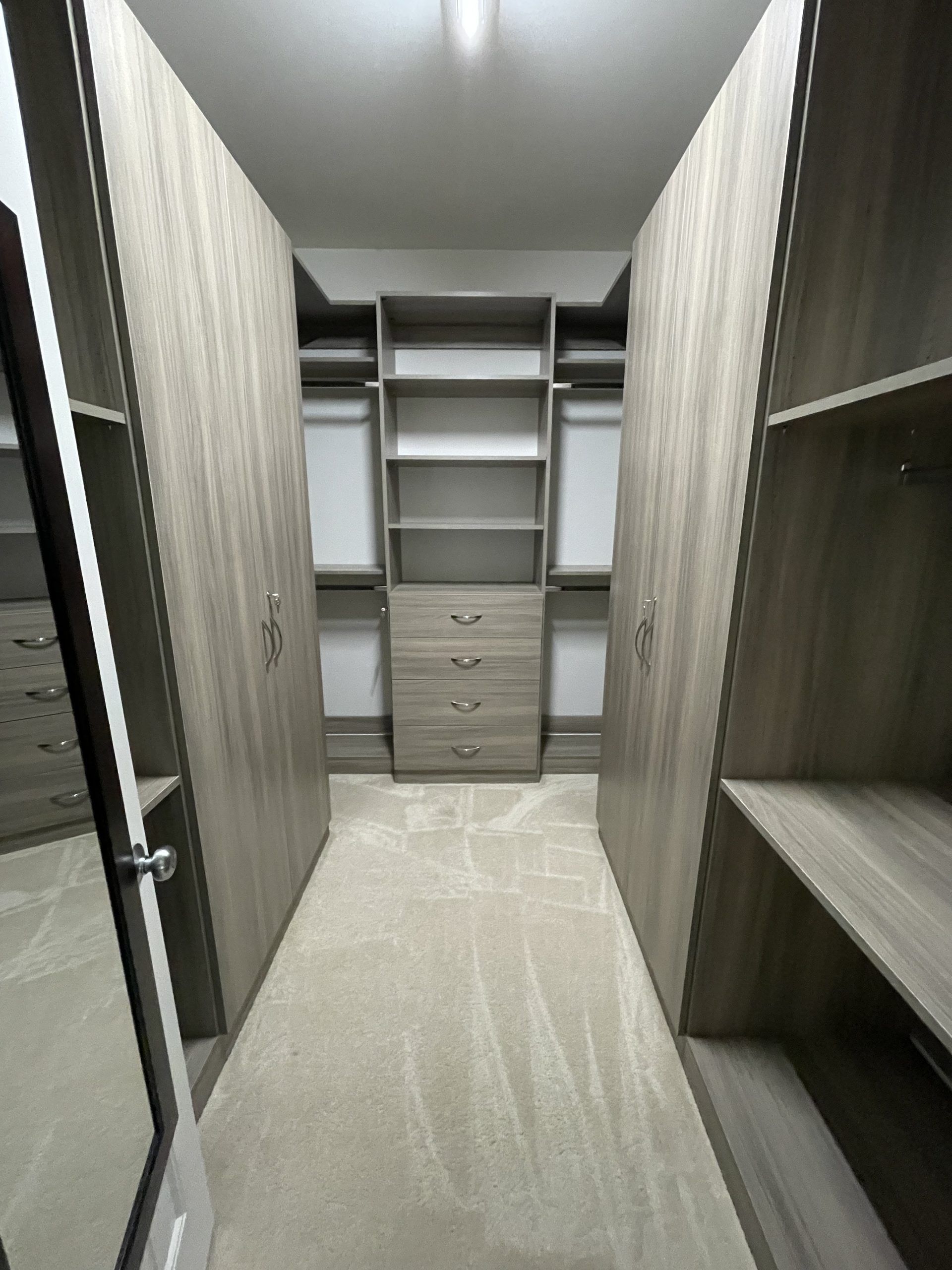 A walk-in closet featuring light brown wood shelving, hanging rods, and a four-drawer dresser on light beige carpet.