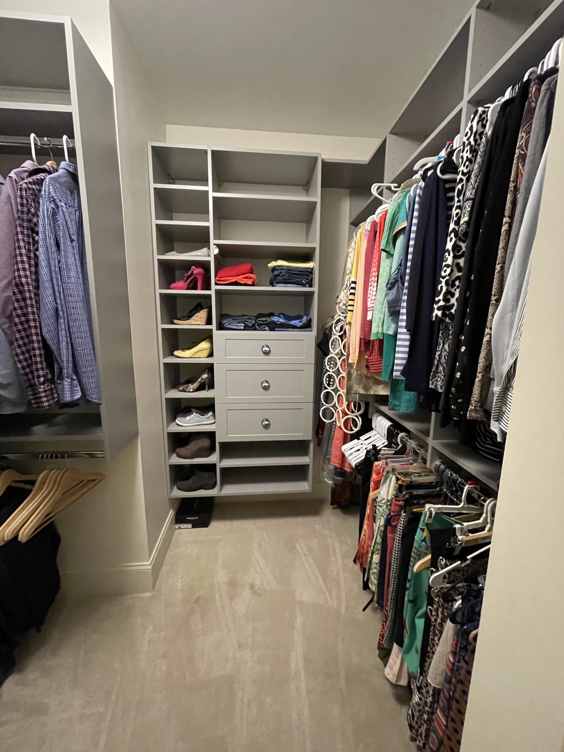 A walk-in closet with gray shelving, hanging clothes on both sides, a center unit with drawers, and carpeted flooring.