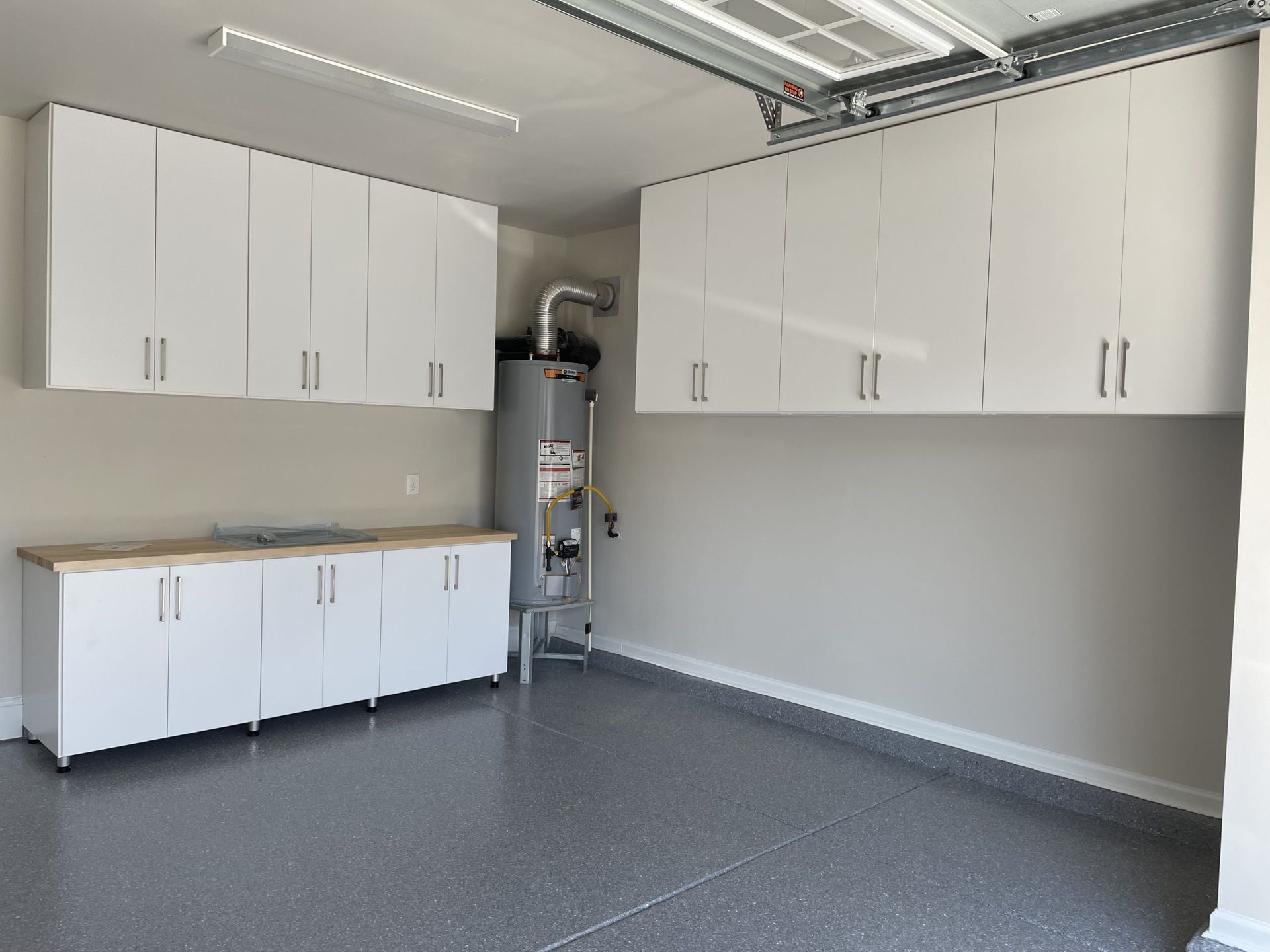 A garage interior featuring white upper and lower cabinetry, a wooden countertop, a water heater, and speckled gray flooring.