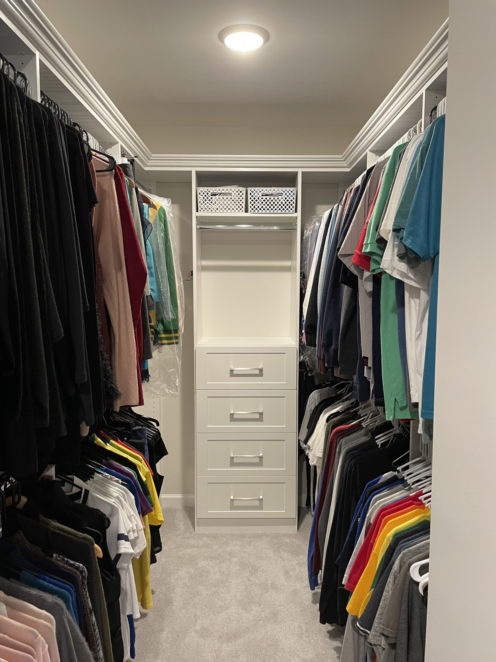 A walk-in closet with hanging clothes on both sides and a central white shelving unit with four drawers and two bins.