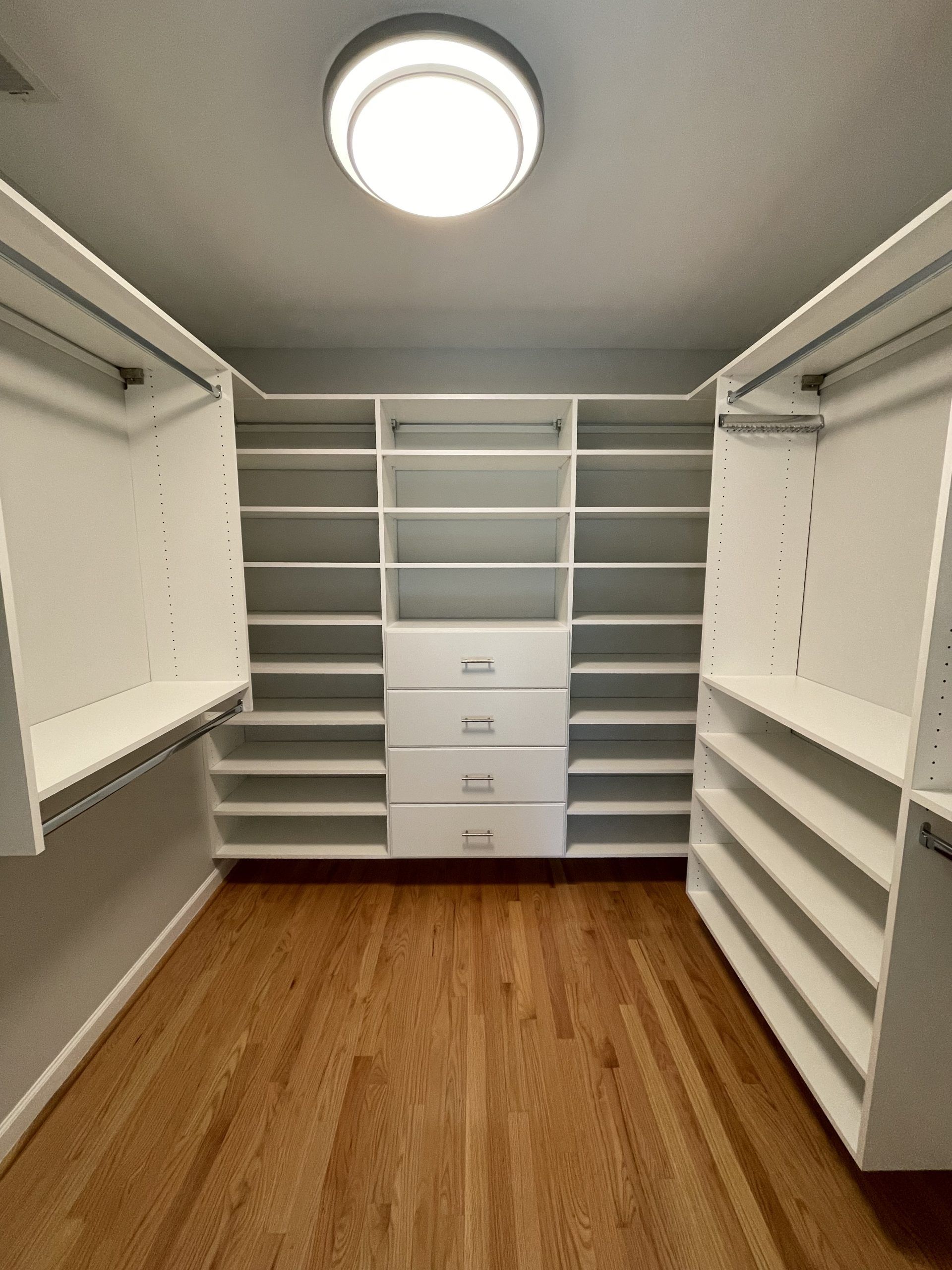 Walk-in closet with white shelving, hanging rods, and a stack of four drawers on hardwood floors under a ceiling light.