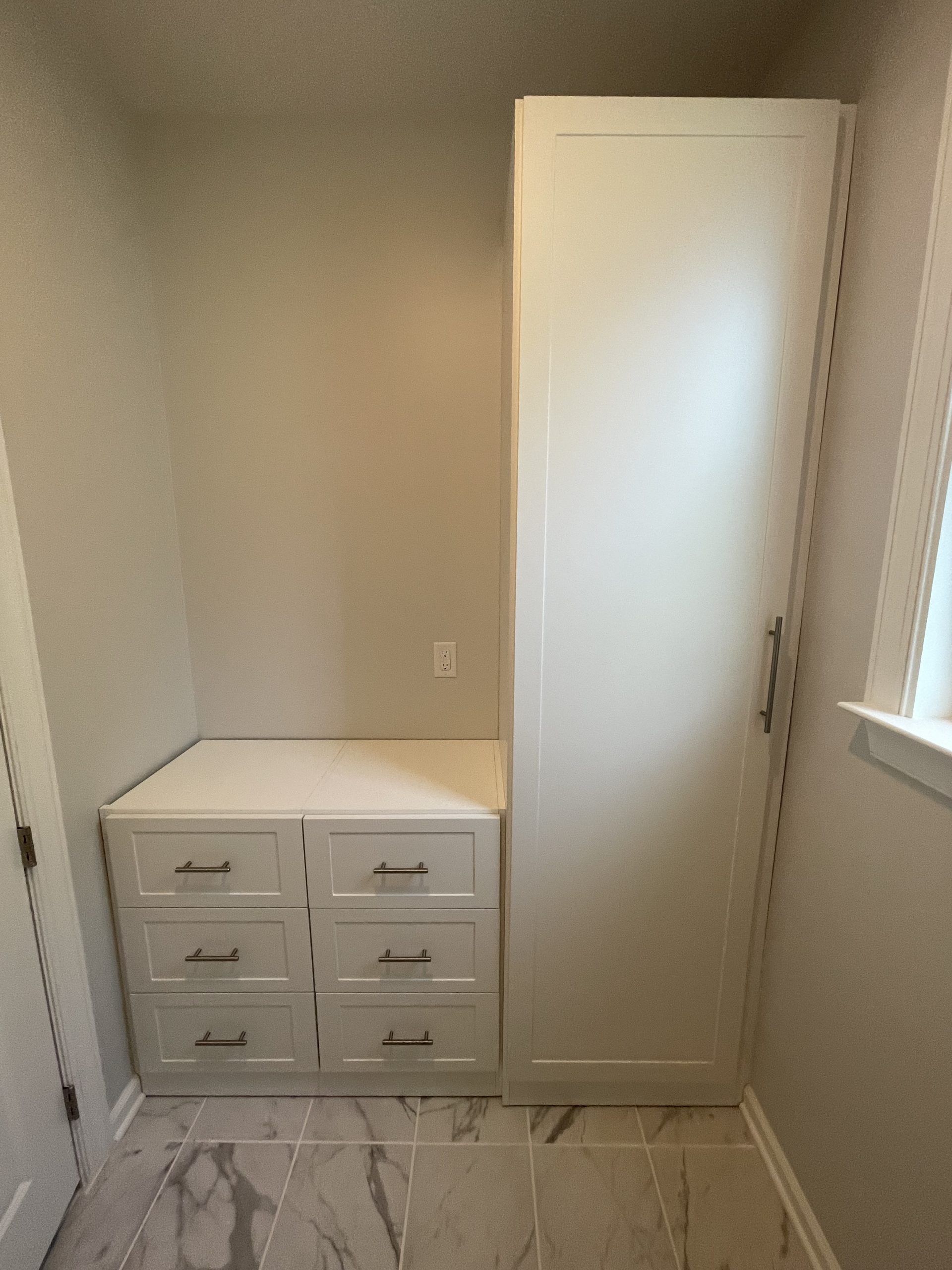 A white, floor-to-ceiling cabinet next to a built-in base unit with six drawers in a room with light-colored tile flooring.
