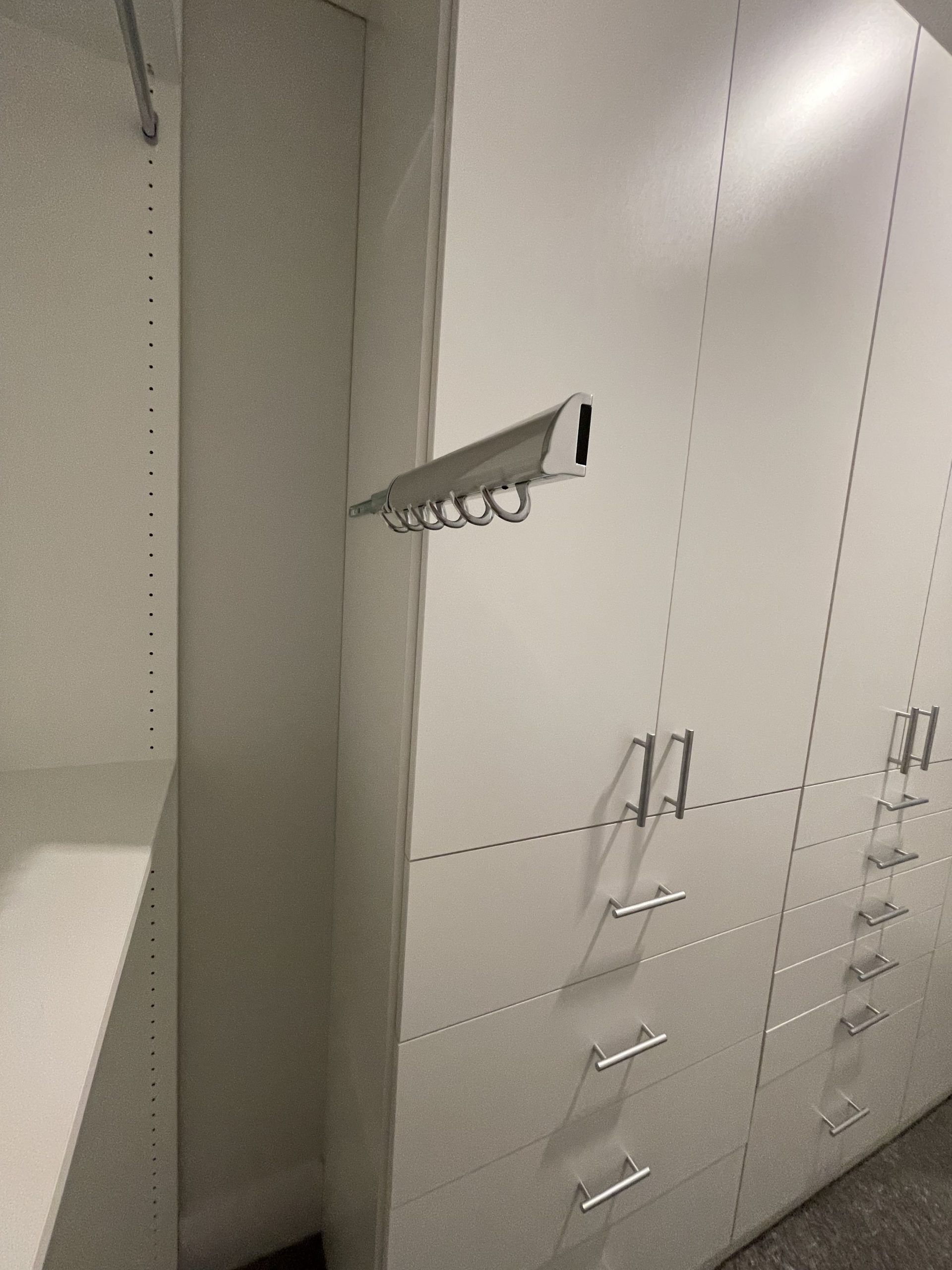 A metal hook rack mounted on the white side panel of a bedroom wardrobe cabinet next to drawers.