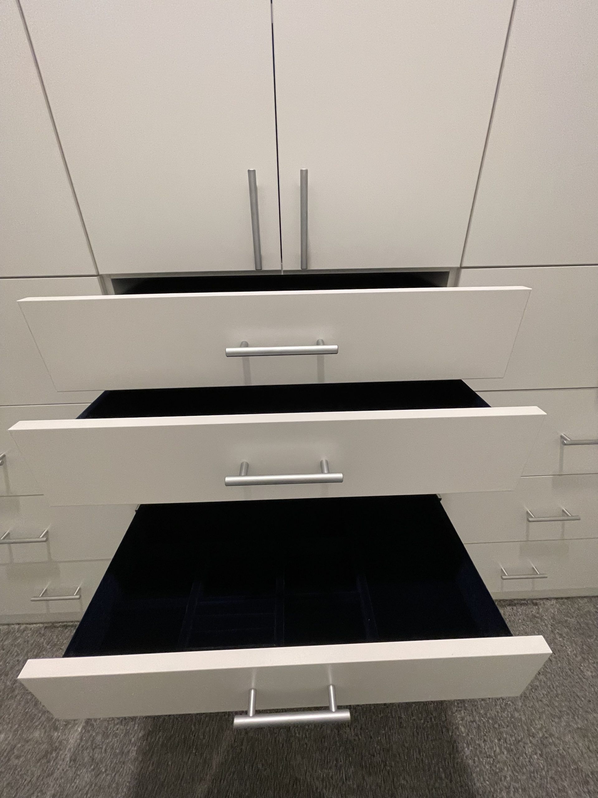 A white closet unit with three drawers pulled open, revealing dark blue velvet-lined jewelry trays inside.
