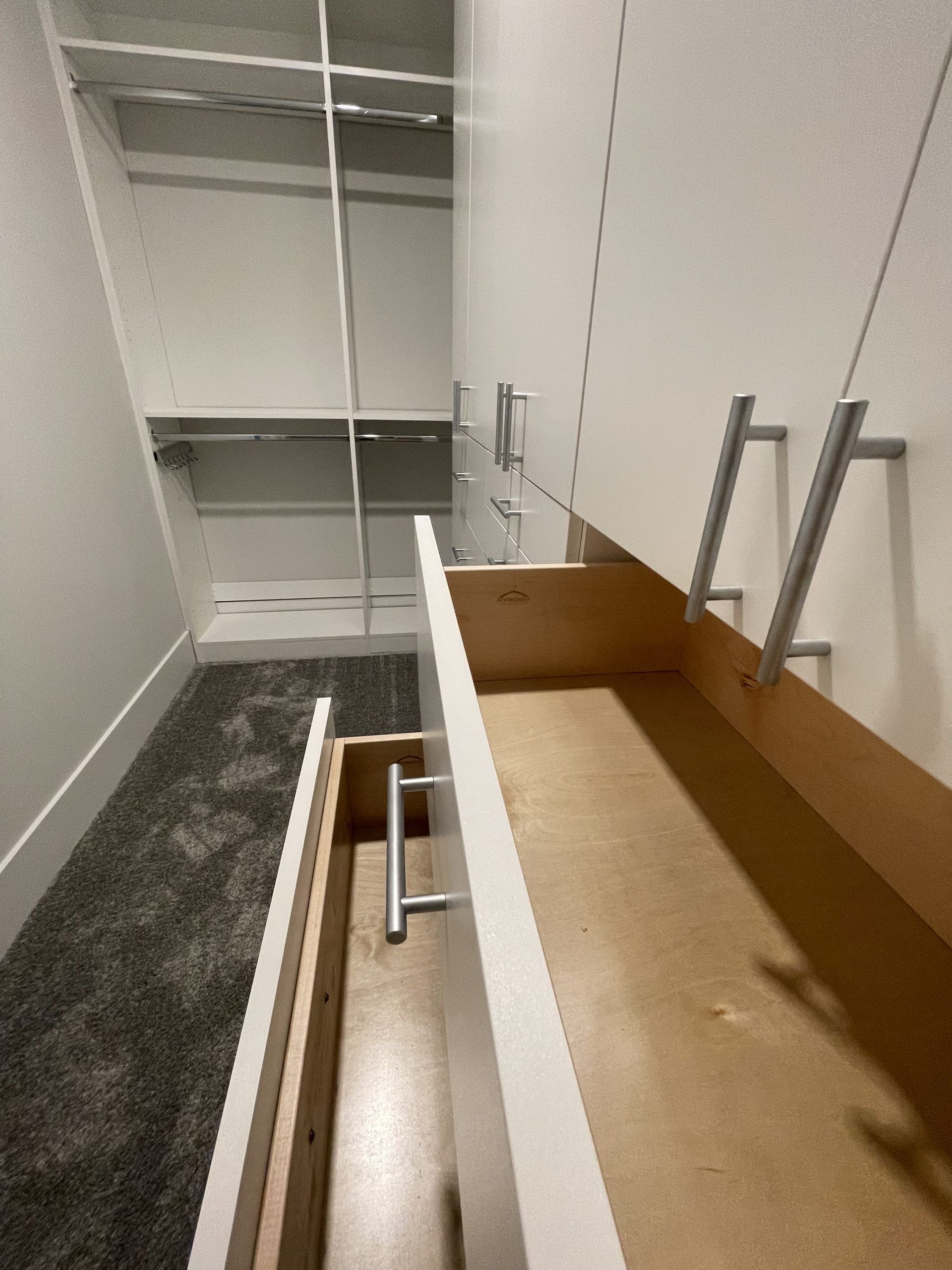 An open, empty wooden drawer in a white closet unit with a gray carpeted floor visible in the background.