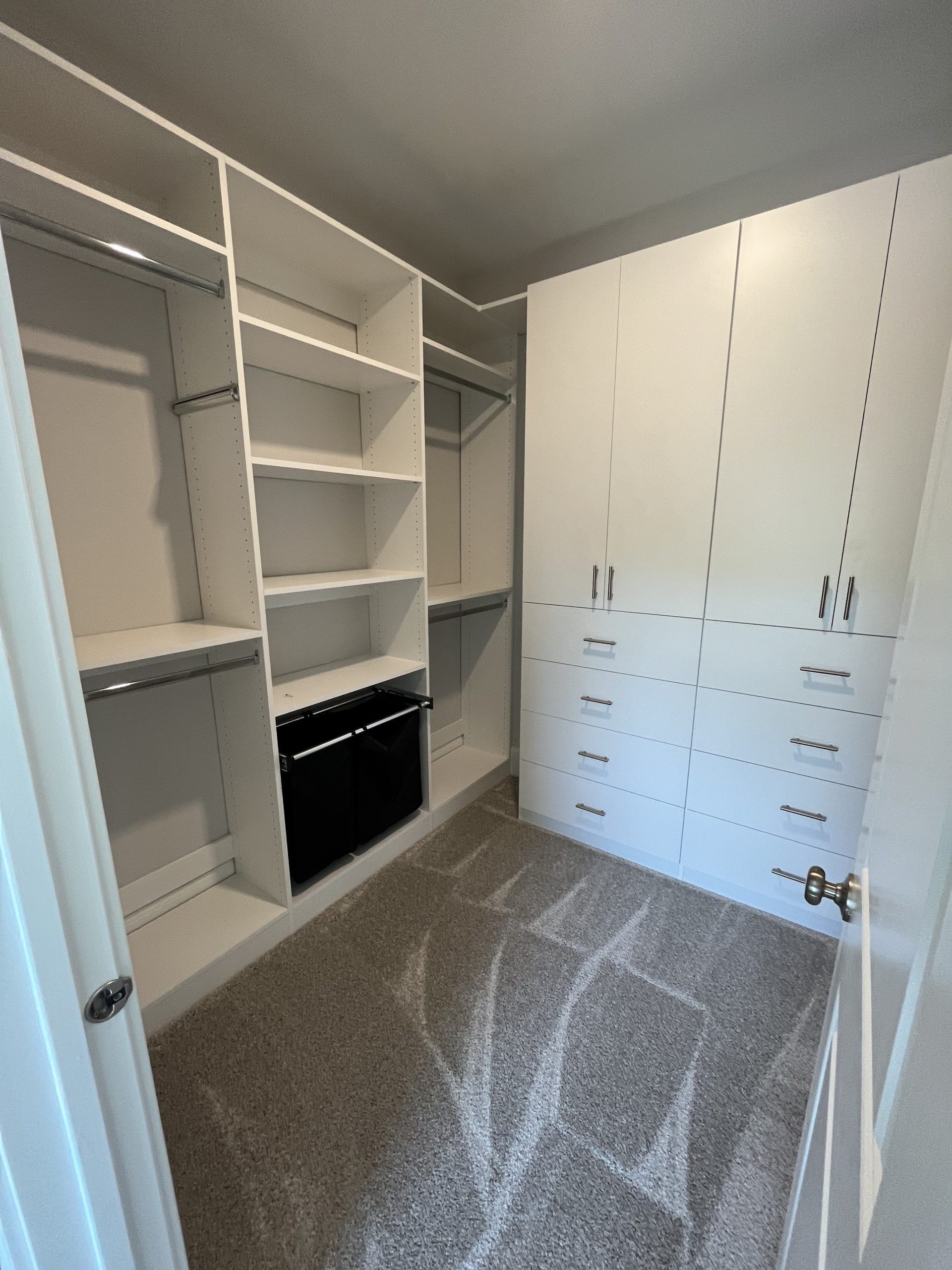 A walk-in closet featuring white shelves, hanging rods, drawers, and a black storage unit on gray carpeted flooring.