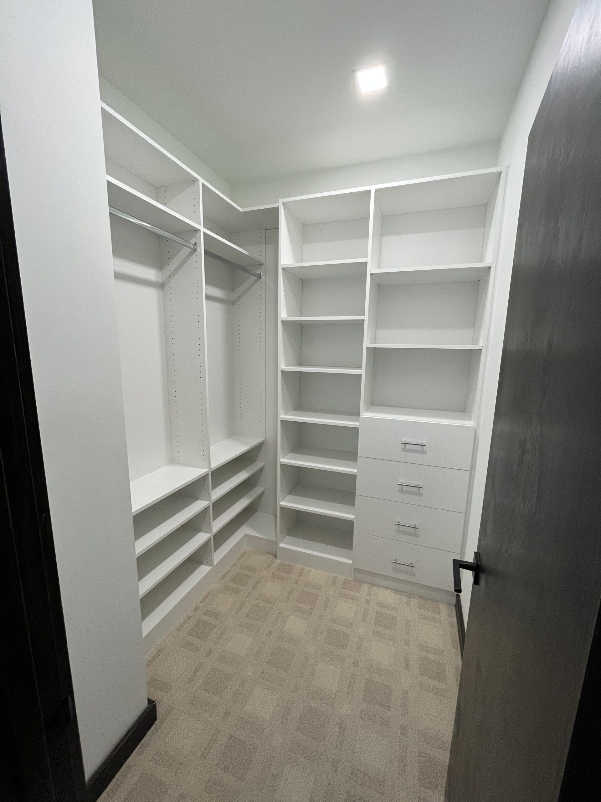A walk-in closet featuring white wooden shelving, clothing rods, and built-in drawers over beige carpet.