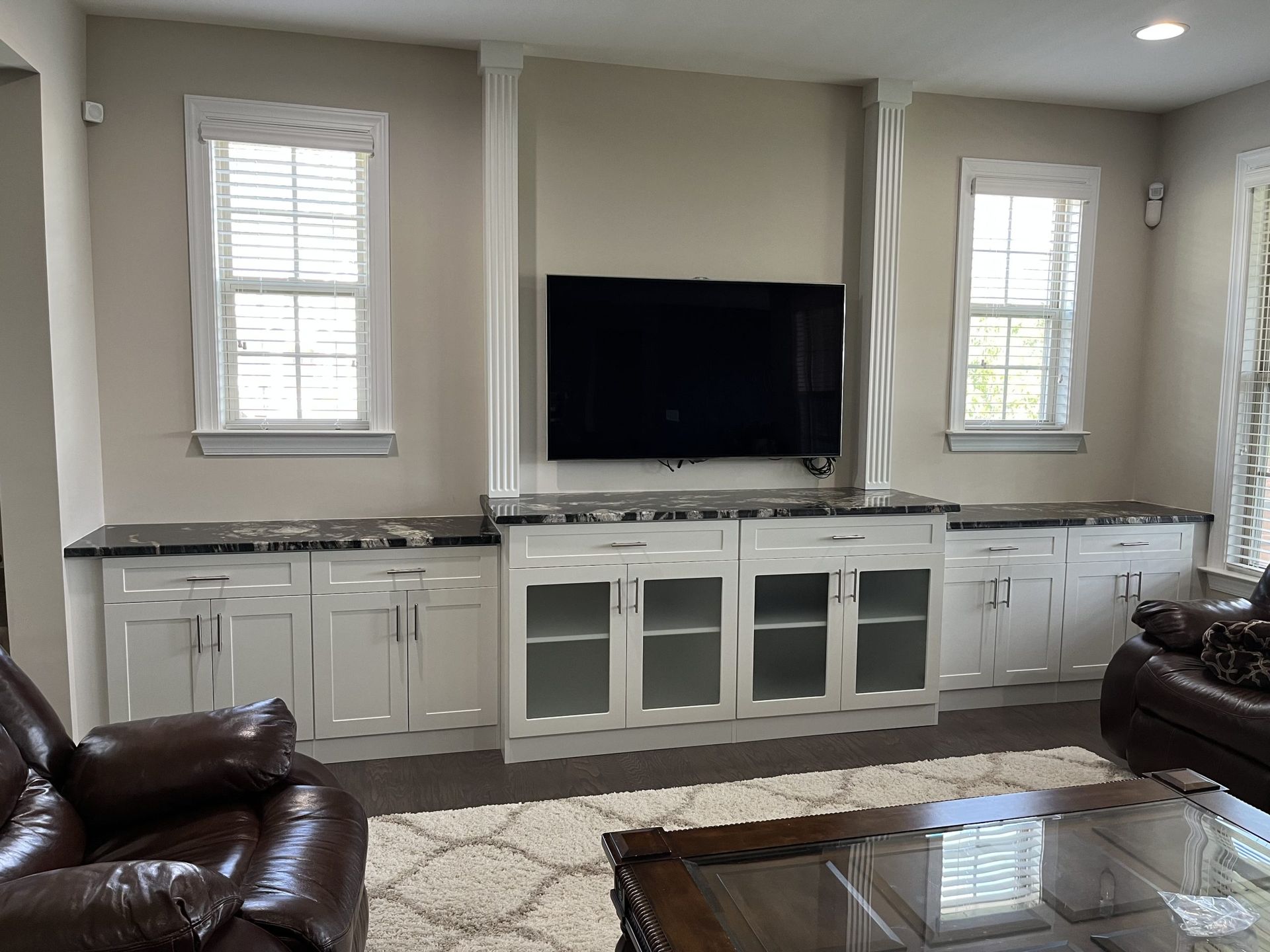 A living room featuring white built-in cabinets with dark countertops, a wall-mounted TV, two windows, and brown furniture.