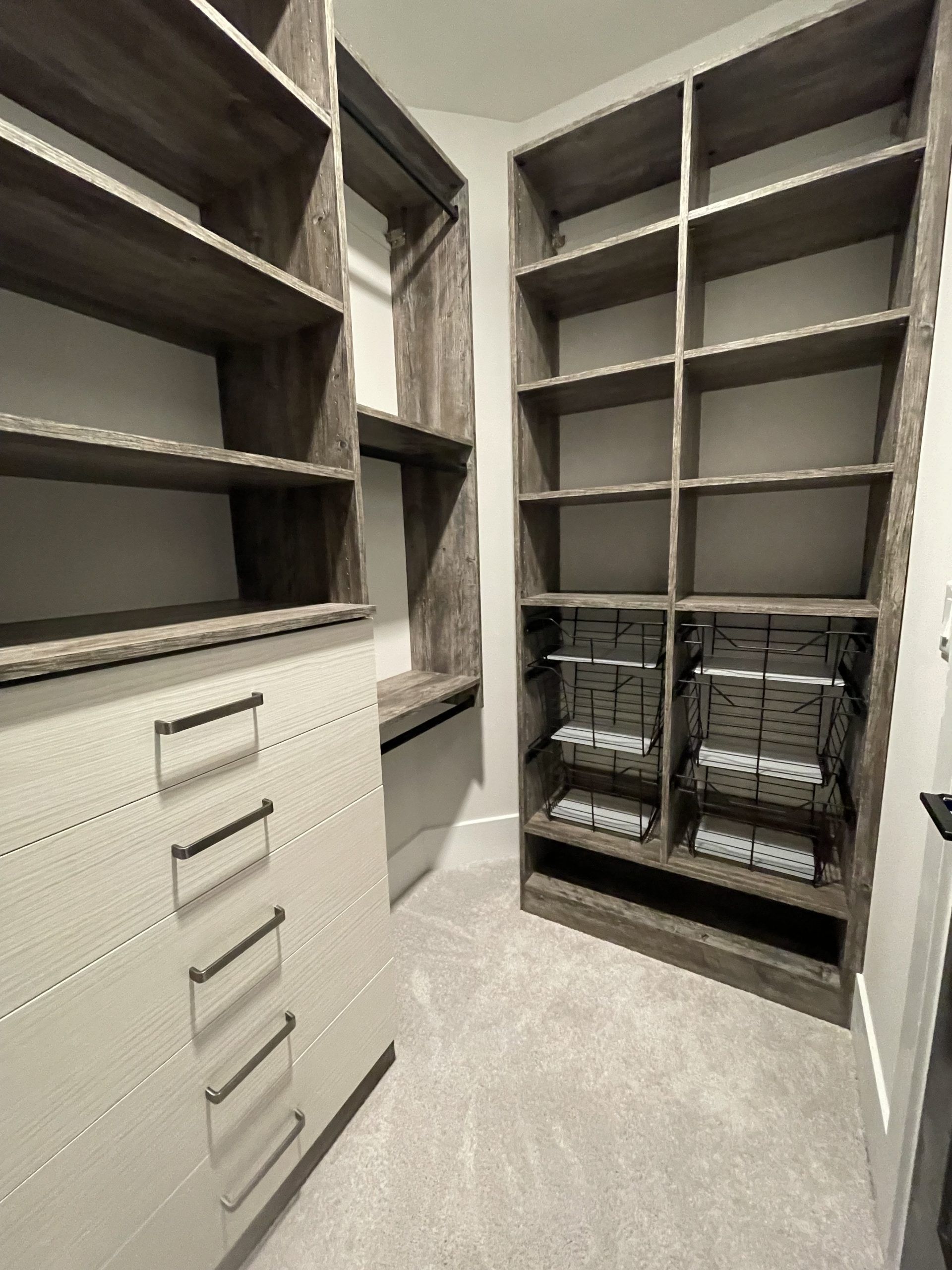 A walk-in closet featuring tall, dark wood shelving units with open compartments and a white drawer stack on the left.