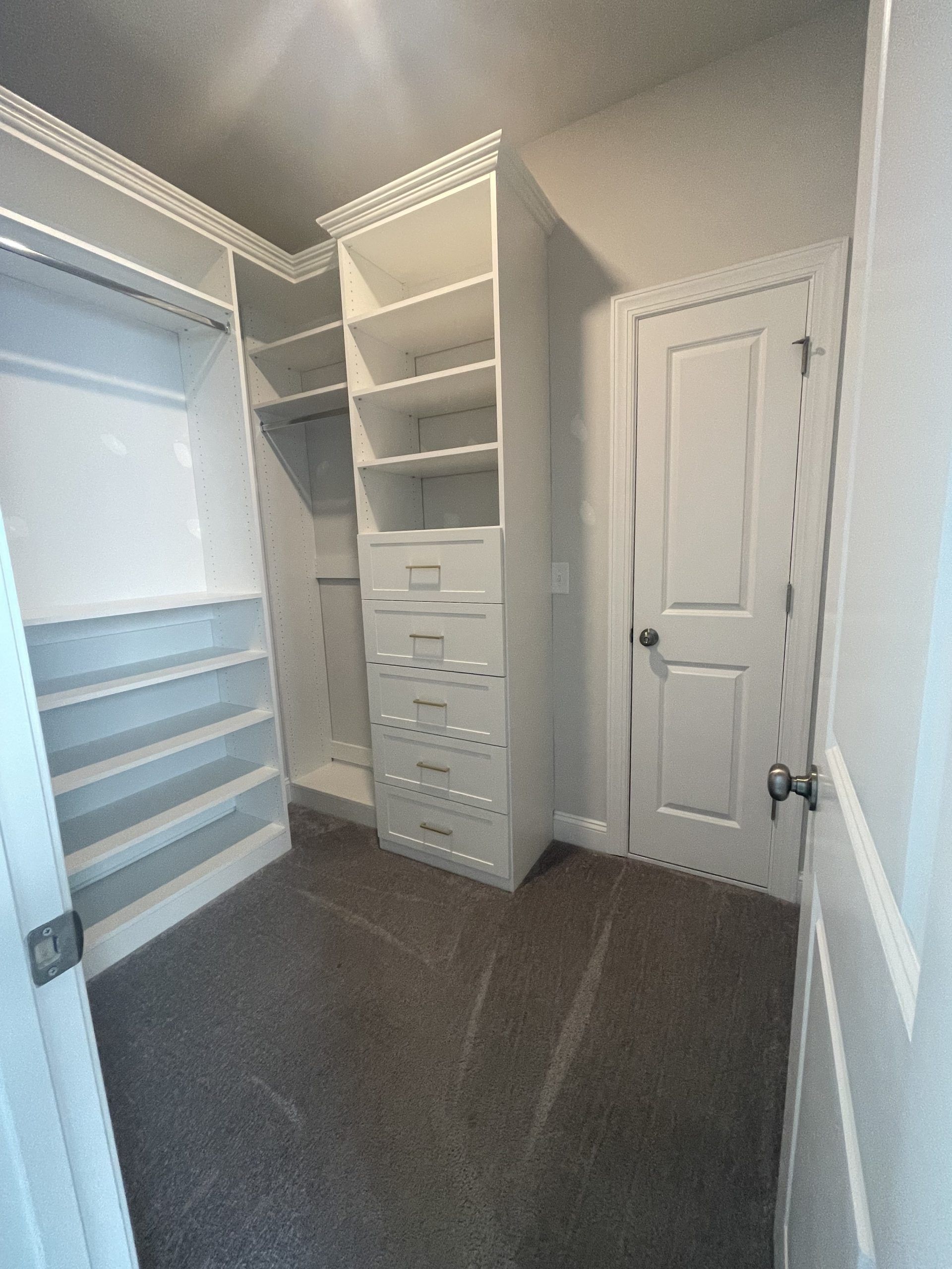 A walk-in closet featuring white wooden shelving, a built-in dresser with drawers, a hanging rod, and gray carpeting.