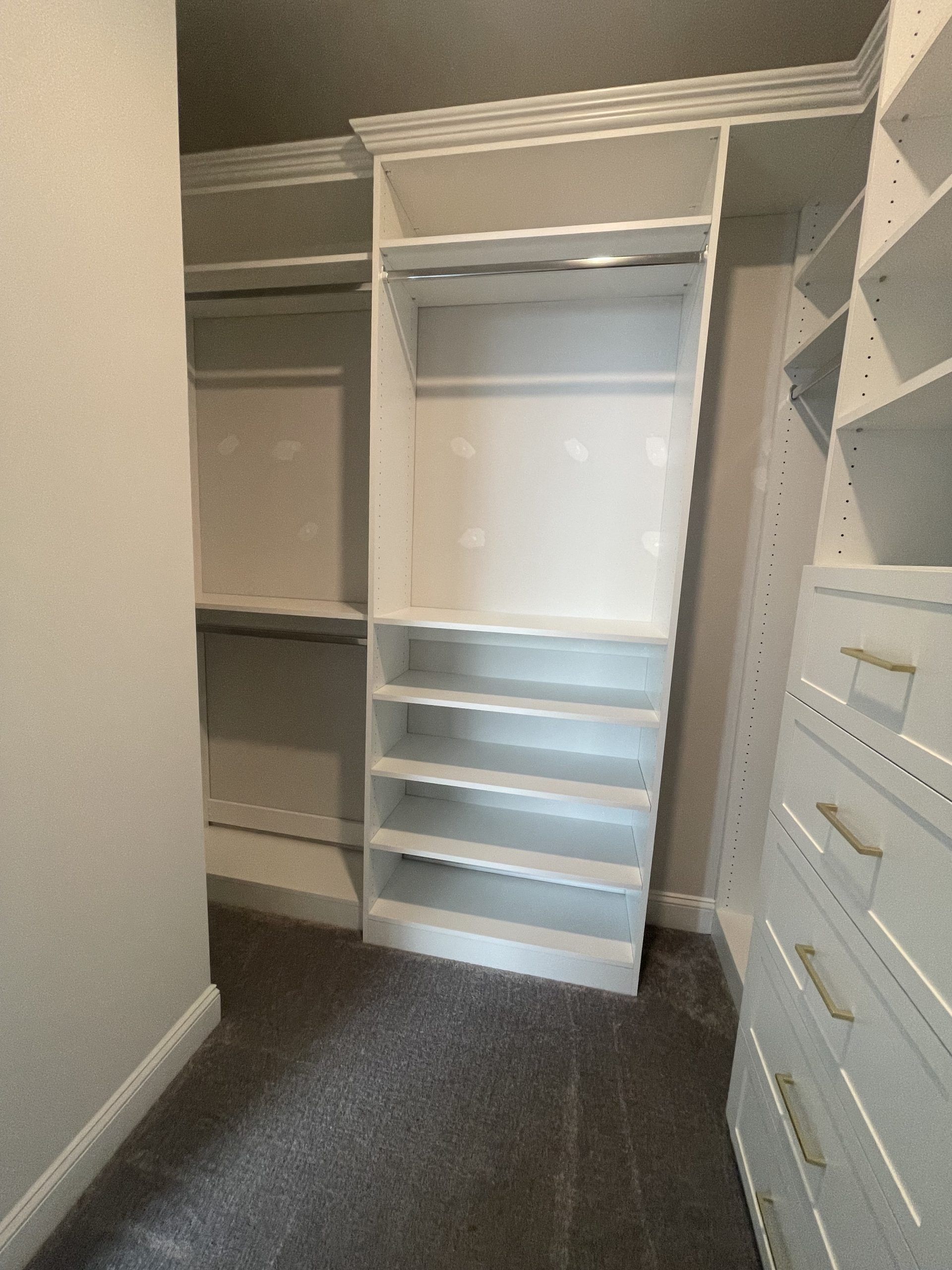 A walk-in closet featuring white wooden shelving, drawers, and hanging rods over gray carpet.