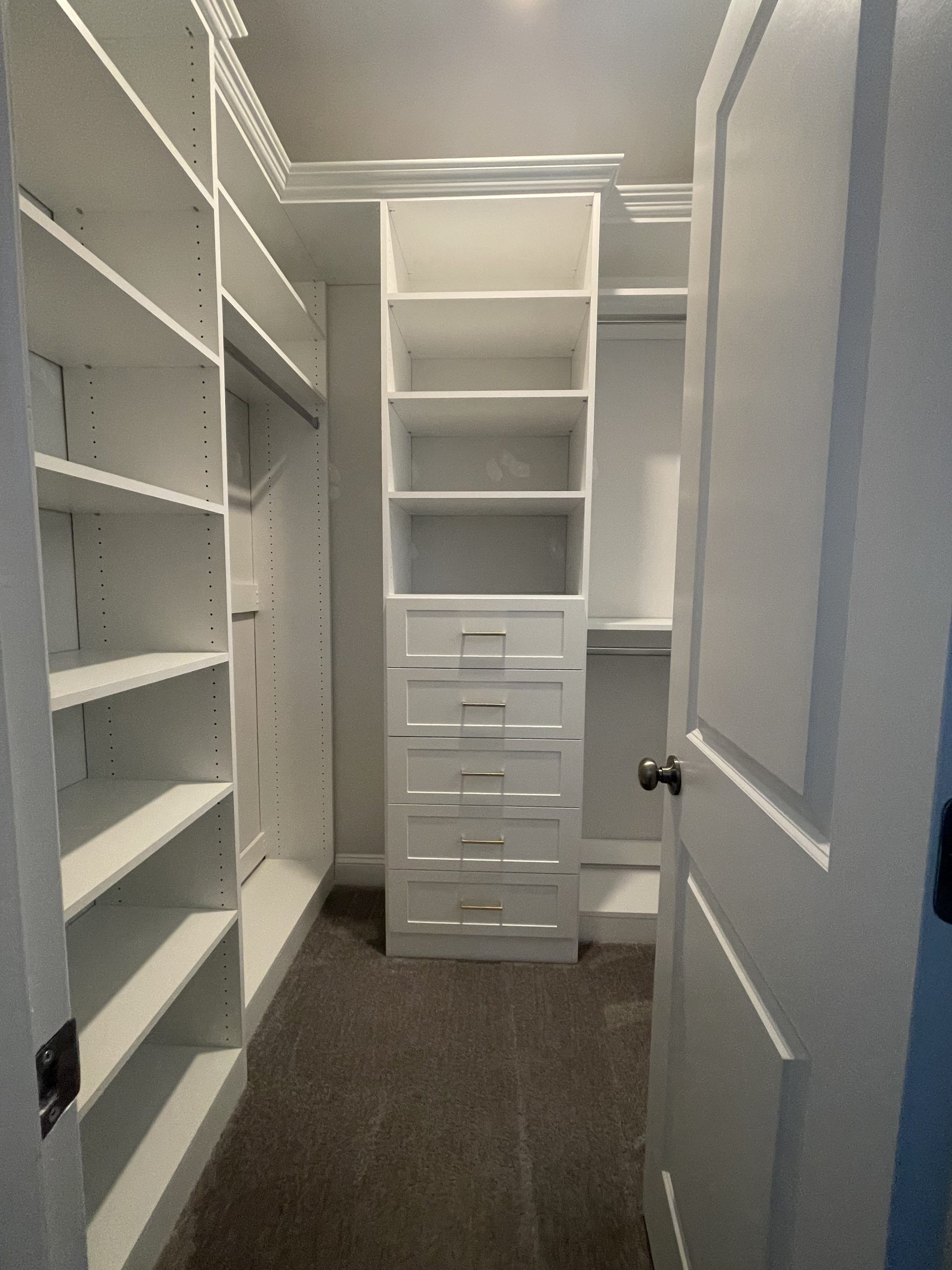 A walk-in closet with white shelving, a central column of drawers, and gray carpeting, seen through an open doorway.