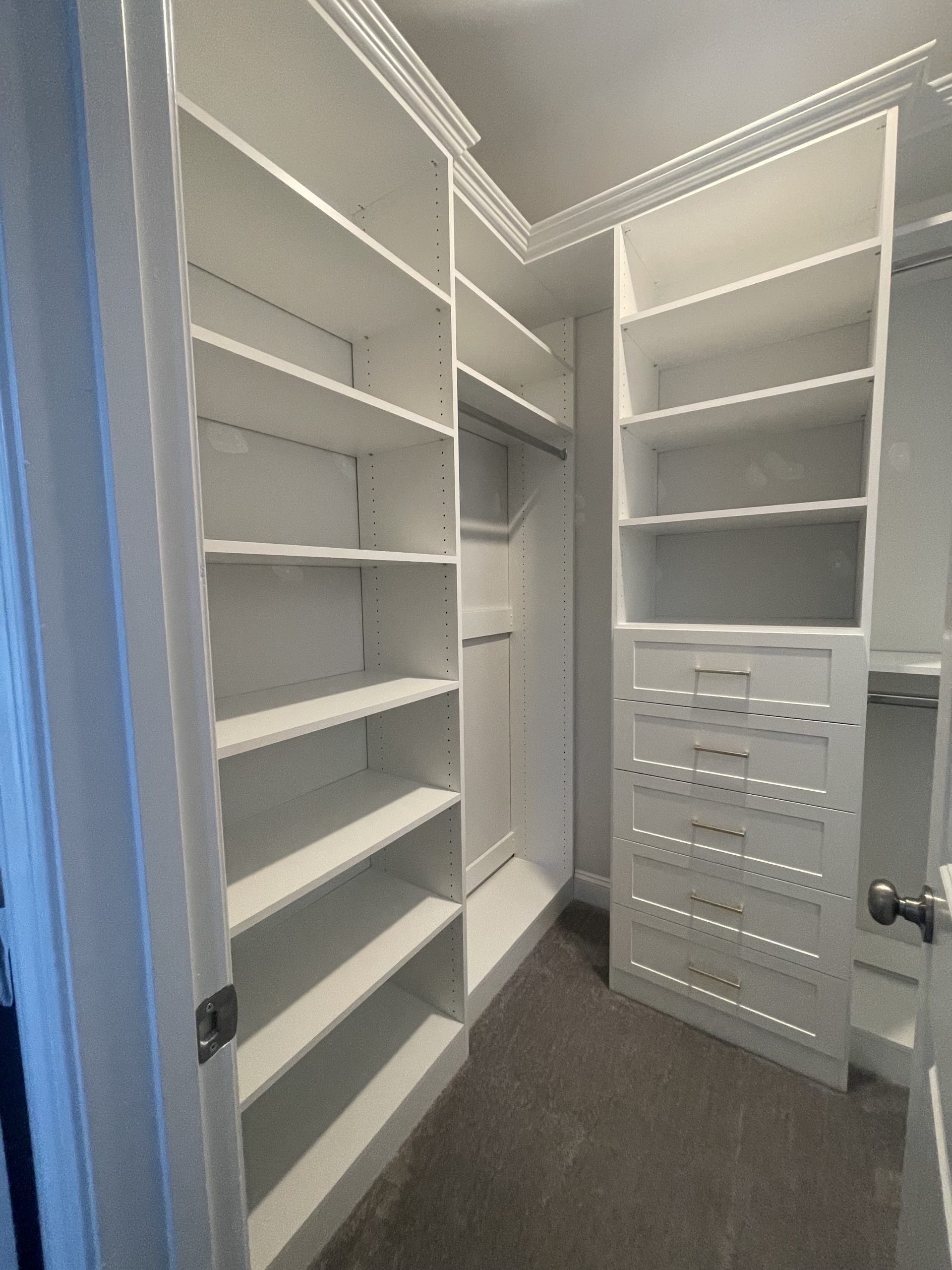 A walk-in closet featuring white wooden shelving, built-in drawers, and a hanging rod on grey carpet.