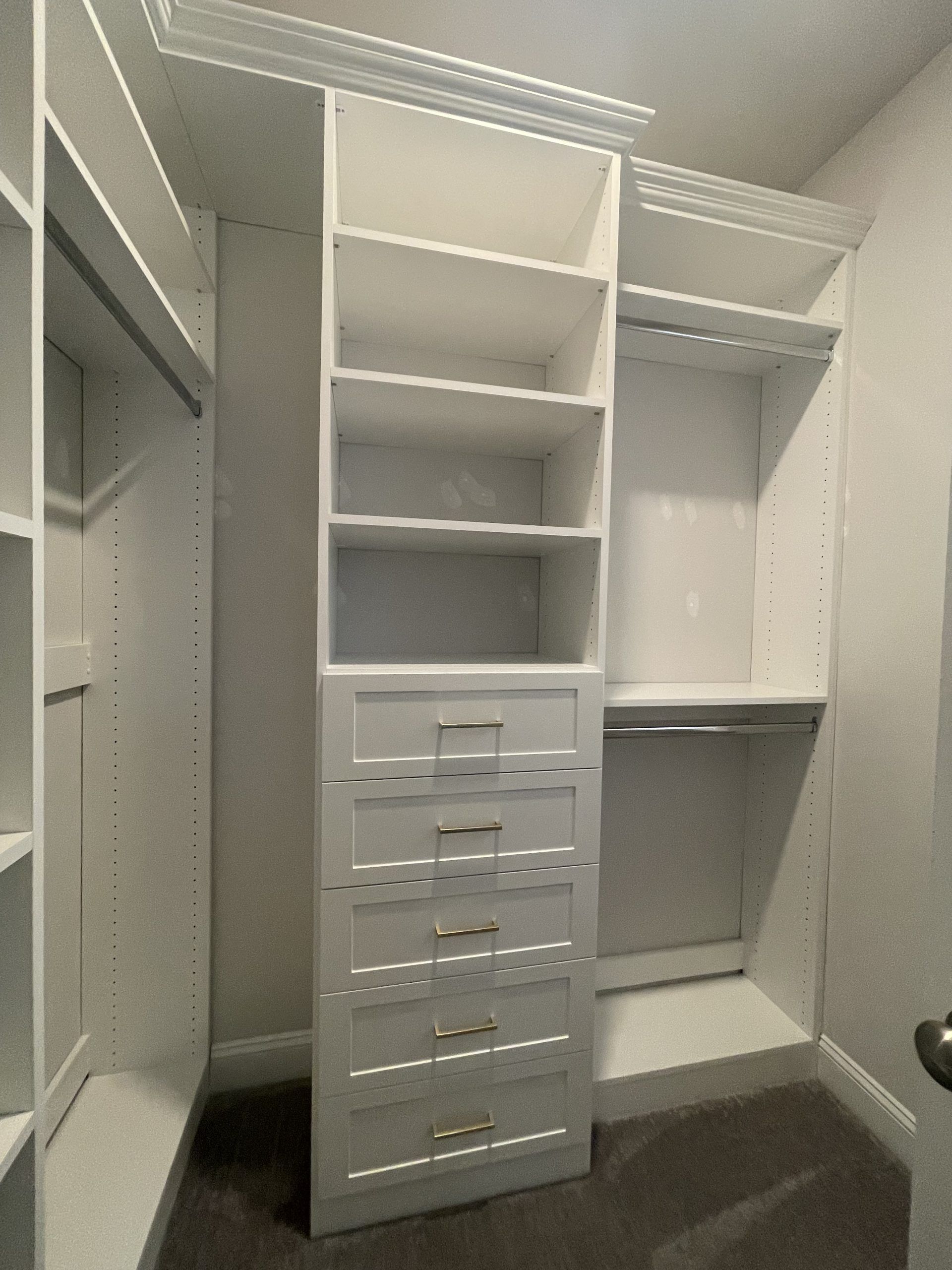 A white custom closet organizer with four open shelves above five stacked drawers with gold pulls, set in a room.