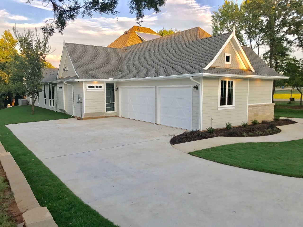 Beige and gray house with a driveway and garage doors, set against a green lawn and blue sky.