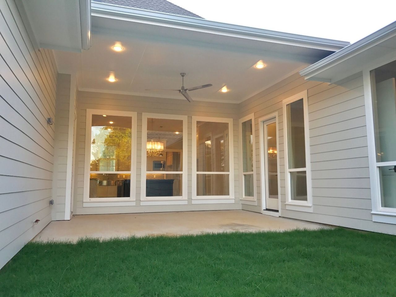 Patio with large windows, recessed lights, and a ceiling fan, overlooking green grass.