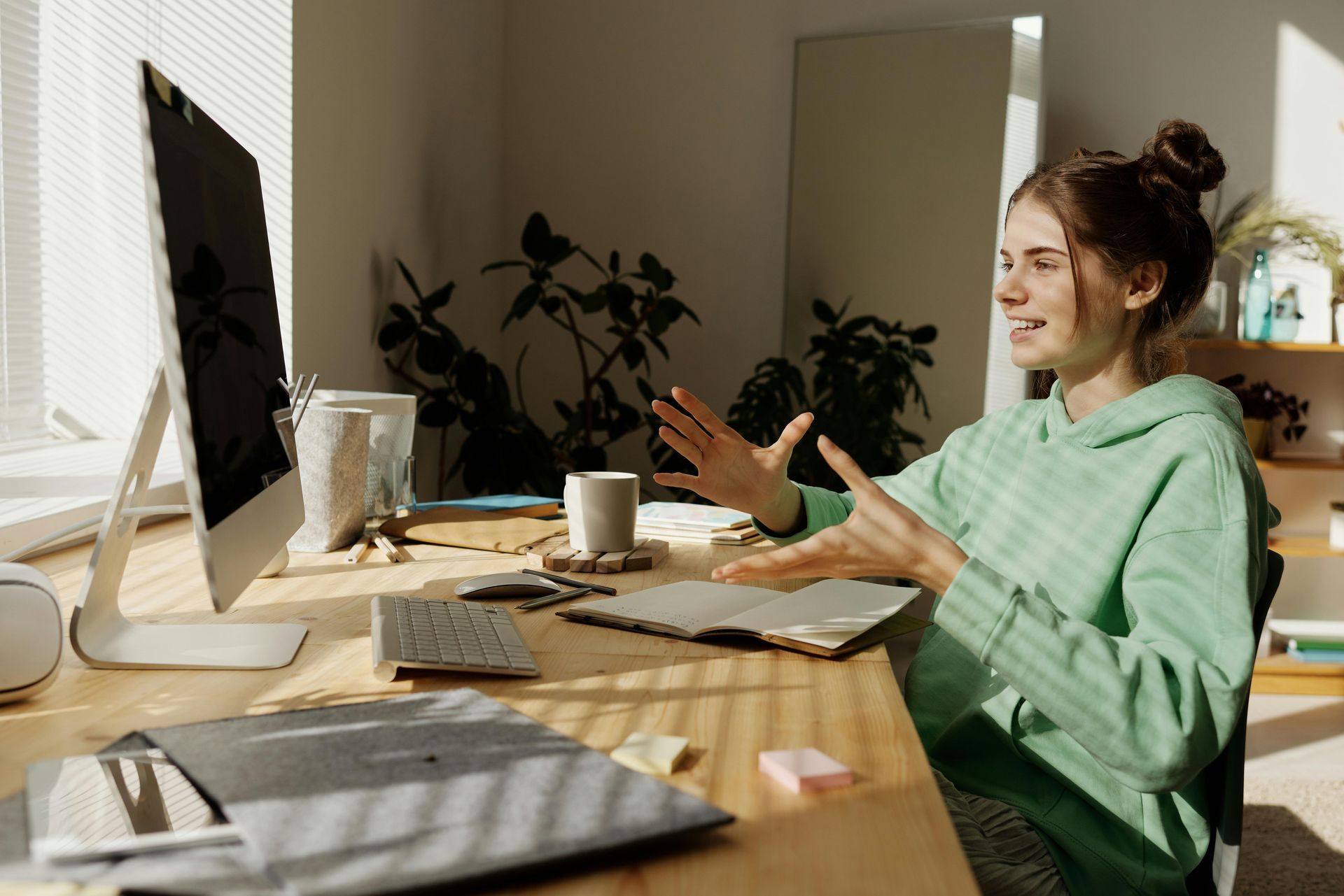 Woman in green hoodie gestures enthusiastically while on a video call at a desk with a computer and notebook.