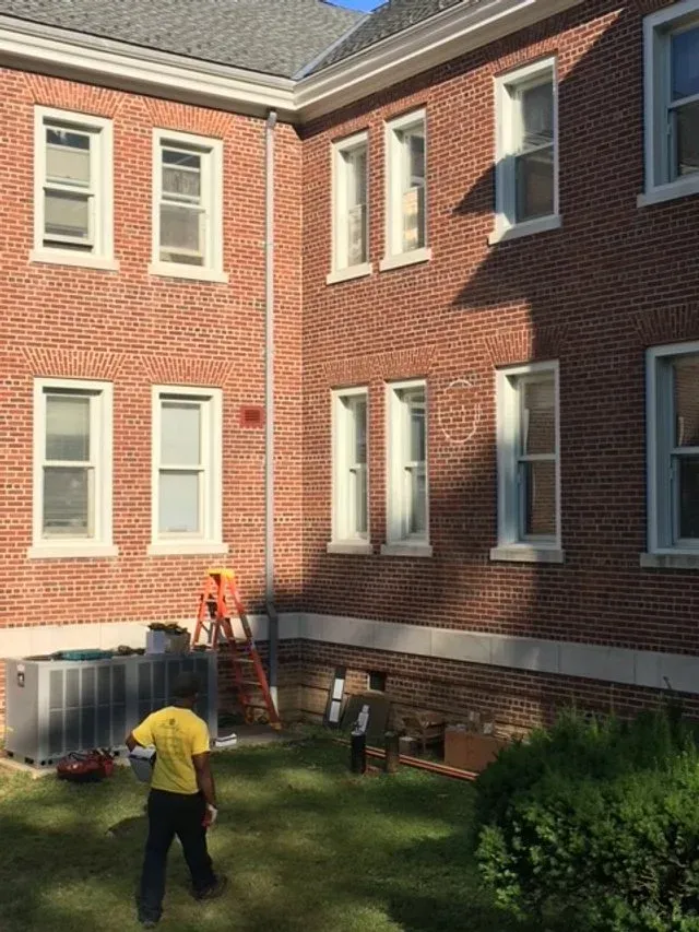 Workers near a red brick building with windows and air-conditioning units in a grassy courtyard