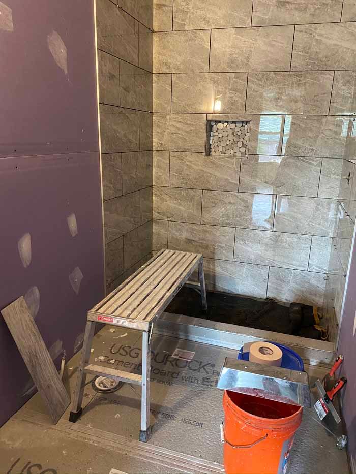 Bathroom under renovation with tiled shower area, bench, bucket, and tools on the floor.
