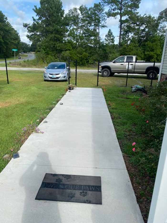 Concrete walkway leading to a street, with a car, pickup truck, grass, and trees in the background.