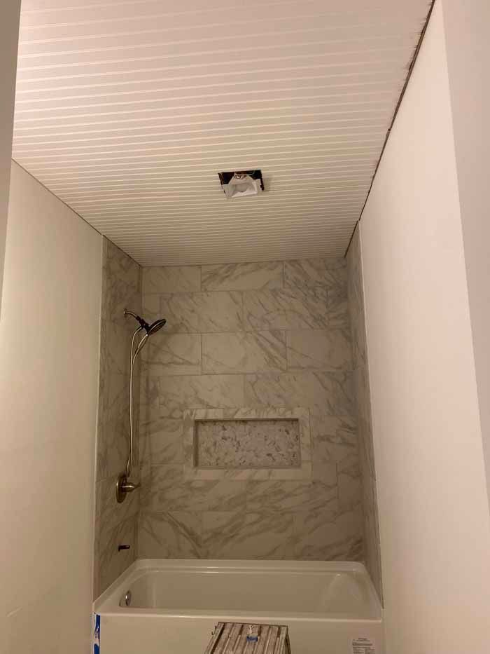 Small tiled bathroom shower with tub, marble-pattern walls, showerhead, and ceiling light.