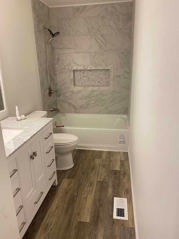 Small bathroom with vanity, toilet, tub, marble shower wall, and wood-look floor; no people involved
