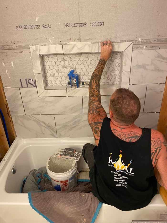 Tile worker kneels in a bathtub, applying marble-look wall tiles in a bathroom under renovation.