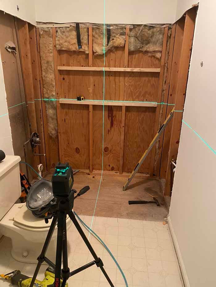 Bathroom under renovation with exposed wall studs, shelves, tools, and a tripod in the foreground