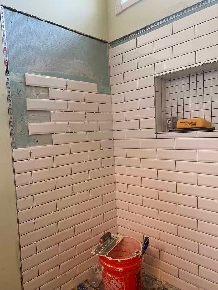 Partially tiled bathroom shower wall with a red bucket, trowel, and a small recessed shelf in progress
