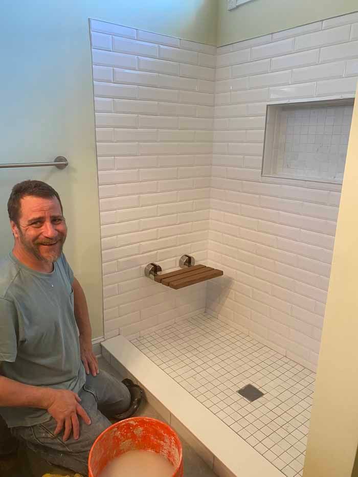 Man sitting beside a white tiled shower under construction, with tools and a bucket nearby