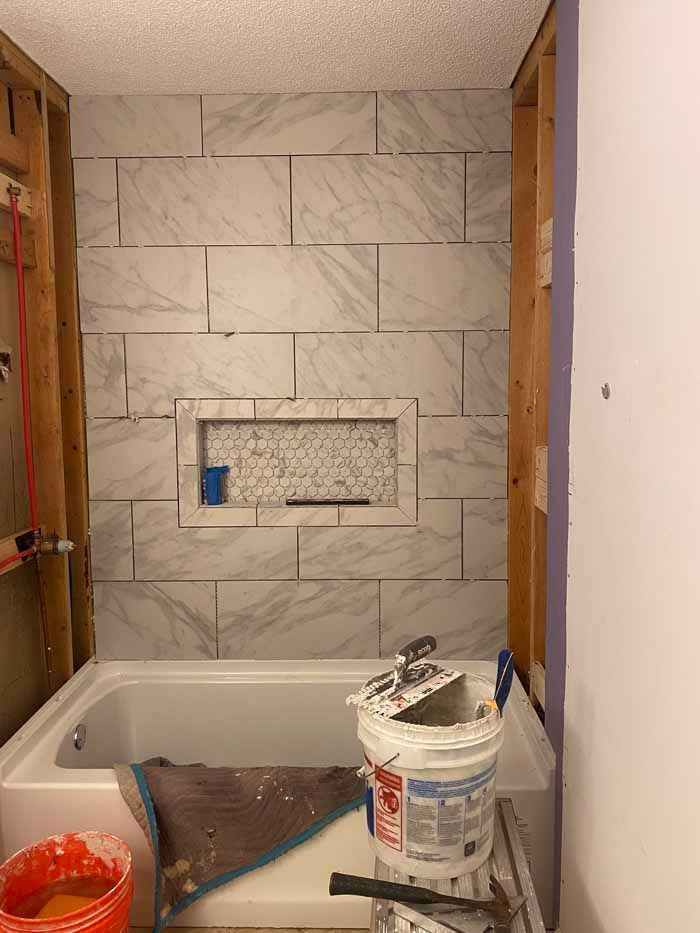 Bathtub alcove under renovation with gray tile wall, tools, and a paint bucket on the tub.