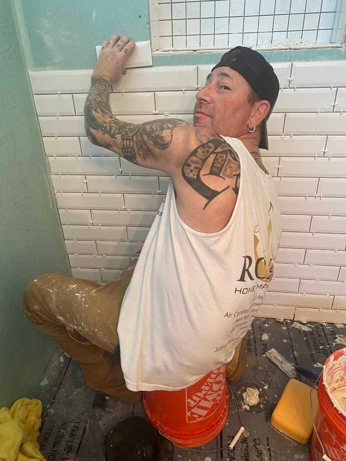 Man tiling a bathroom wall, kneeling on a bucket beside white subway tiles and blue trim.