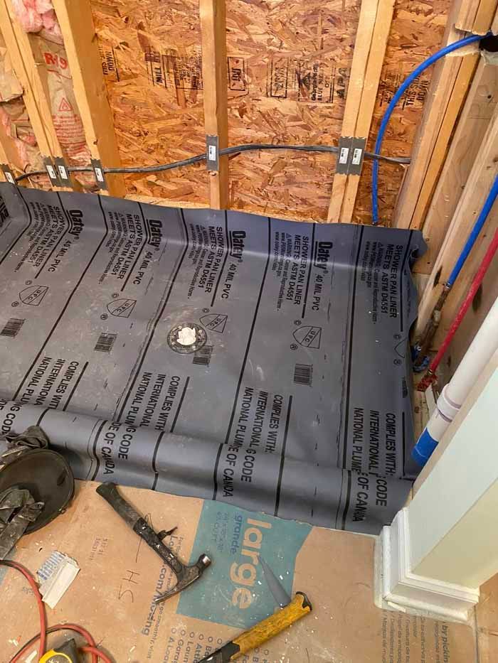 Partially installed floor membrane over subfloor in a construction site, with exposed wall framing and blue tubing.
