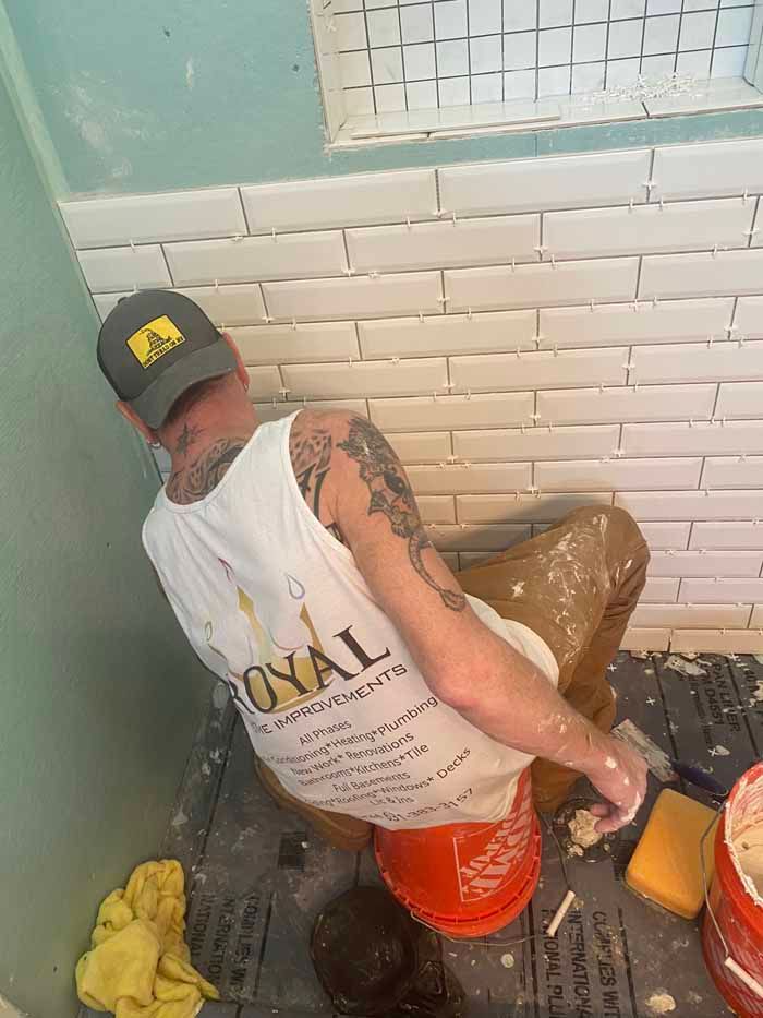 Person cleaning a tiled bathroom while holding a bucket and tool, wearing a cap and white tank top.